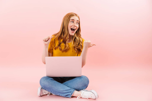 Emocionální úsměv veselá dívka izolované přes růžové stěny pozadí pomocí notebooku ukazující cpyspace. - Fotografie, Obrázek