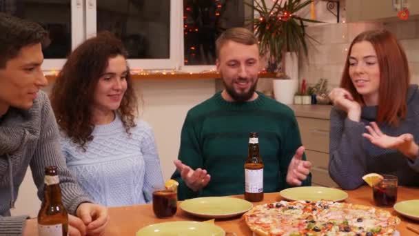 お祝いのテーブルに座って新年を祝う幸せな友人は自宅でパーティーを前夜。男と女はピザのスライスを取る.テーブルの上にビールとマルチワイン。休日やお祝いの概念。4k映像. - 映像、動画