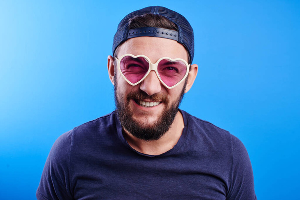 Cheerful bearded Caucasian guy in a cap and girl's glasses smiles joyfully, feels young and happy on a blue studio background - Foto, immagini