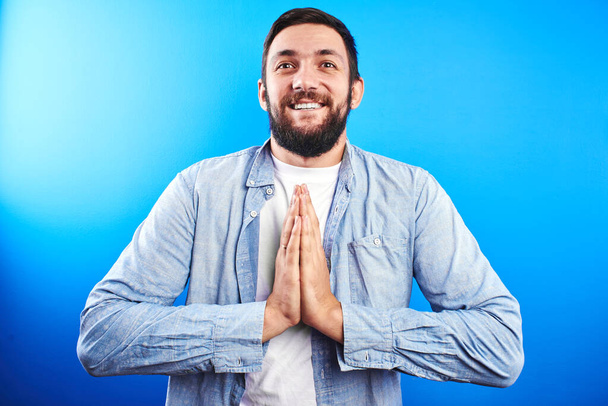 Дружелюбный добрый кавказский бородатый мужчина улыбается с сложенными в молитве руками благодарственная поза изолирована на синем фоне
 - Фото, изображение