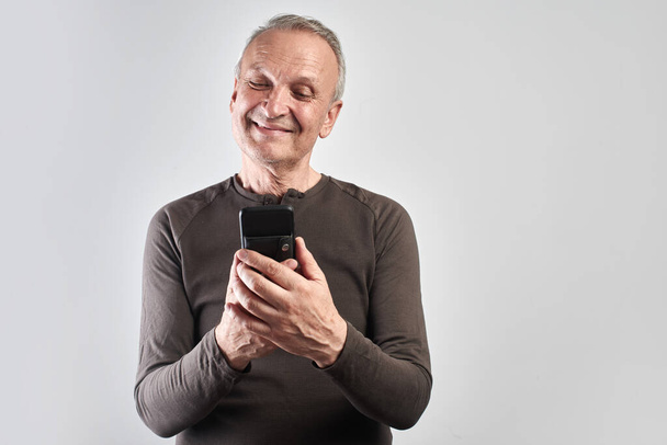 Elderly handsome man with happy face looks at phone screen isolated in white studio, the older generation is mastering high technology - Foto, immagini