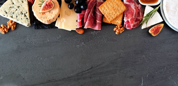 Assorted cheeses and deli meat appetizers. Top border, above view banner on a dark stone background with copy space. - Φωτογραφία, εικόνα