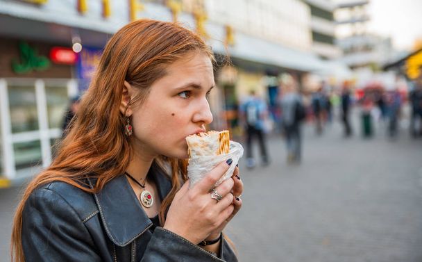 portrait of young teenager redhead girl with long hair eating chicken shawarma on street - Foto, afbeelding