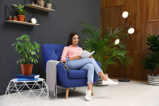 Happy woman reading book in armchair at home. Indoor plants for trendy interior design - Foto, imagen