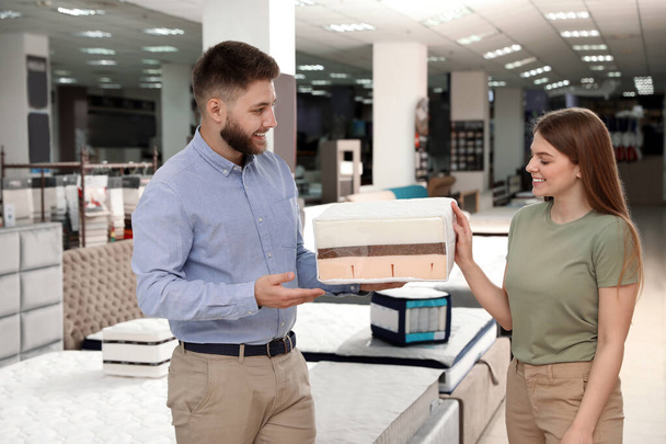 Young salesman showing sample of modern orthopedic mattress to customer in store - Zdjęcie, obraz