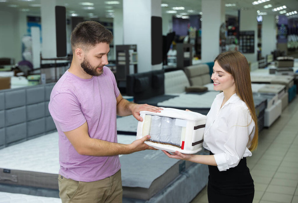 Young saleswoman showing sample of modern orthopedic mattress to customer in store - Zdjęcie, obraz