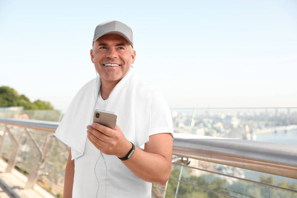 Handsome mature man in sportswear with mobile phone on bridge, space for text. Healthy lifestyle - 写真・画像