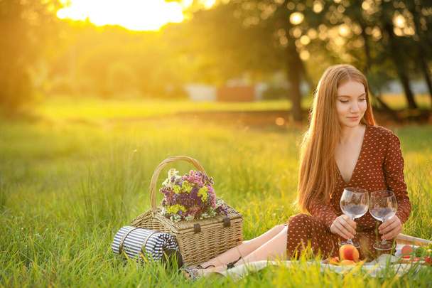 Beautiful young woman with wineglasses and picnic basket sitting on blanket in park - Fotoğraf, Görsel
