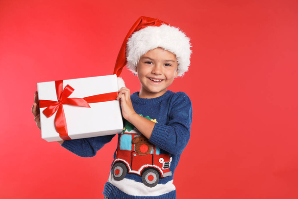 Happy little child in Santa hat with gift box on red background. Christmas celebration - Фото, зображення