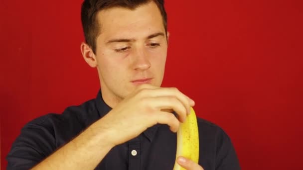 mężczyzna w koszuli czyści i zjada banana na czerwonym tle - Materiał filmowy, wideo