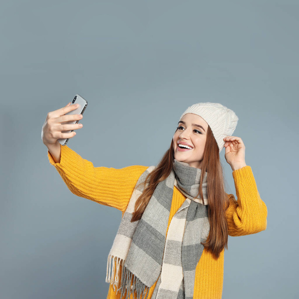 Beautiful young woman in warm clothes  taking selfie on grey background. Winter season - 写真・画像