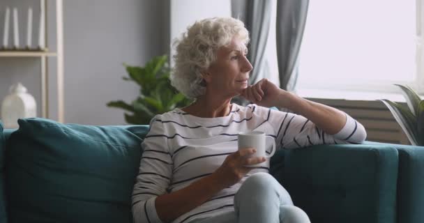Serieuze bedachtzame oude vrouw die weg kijkt drinkt thee op de bank - Video