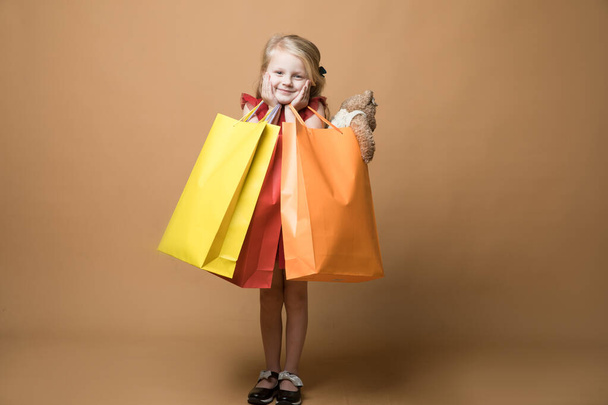 Ένα νεαρό κορίτσι με κόκκινο φόρεμα και τσάντες για ψώνια, πολύ χαρούμενα ψώνια. Ένα νεαρό κορίτσι στέκεται σε πορτοκαλί φόντο. - Φωτογραφία, εικόνα