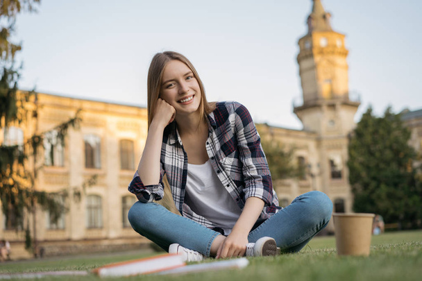 Opiskelijatyttö istuu nurmikolla puistossa. Koulutuskäsite. Muotokuva nuori viehättävä nainen katselee kameraa ja nauraa
  - Valokuva, kuva