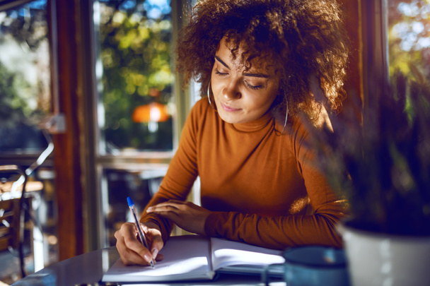 Retrato de un lindo estudiante de raza mixta con pelo rizado y cuello alto sentado en la cafetería y estudiando para los exámenes
. - Foto, Imagen