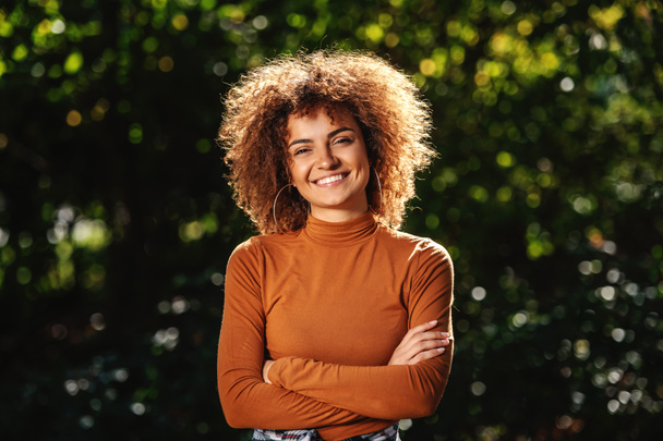 Retrato de la hermosa mujer joven de raza mixta sonriente con el pelo rizado de pie en el parque con los brazos cruzados
. - Foto, Imagen