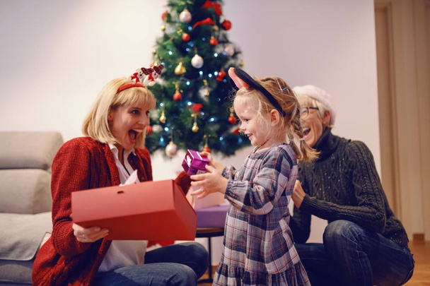 verblüffte junge Mutter mit Weihnachtsgeschenk von ihrer liebenden Tochter. Im Hintergrund Tannenbaum und Großmutter. - Foto, Bild