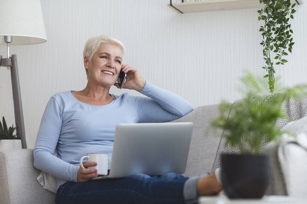 Aktiivinen vanha nainen pitää kannettavaa tietokonetta ja puhuu älypuhelimella
 - Valokuva, kuva