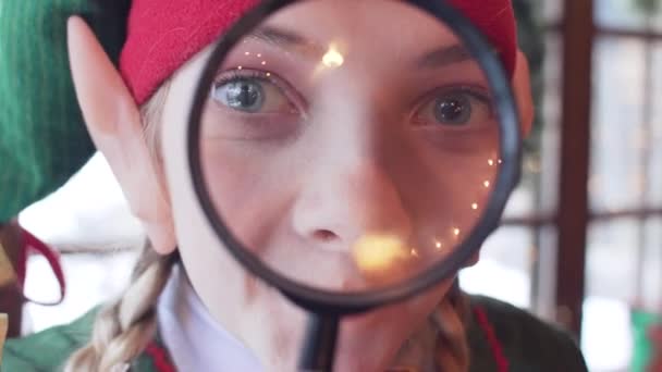 Cheerful elf girl looking at the camera through a magnifying glass. close-up - Πλάνα, βίντεο
