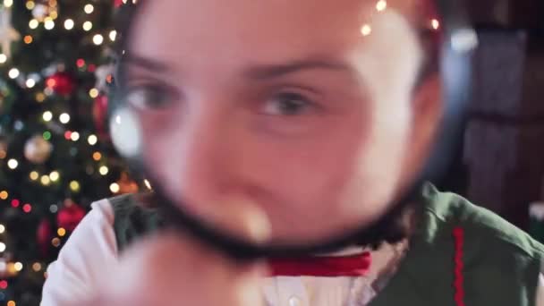 Elf looks into the camera through a magnifying glass, showing teeth. Christmas tree in the background. Close-up - Footage, Video