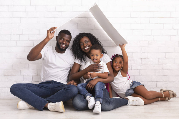 Familia afroamericana feliz sentada en el suelo bajo el techo simbólico
 - Foto, imagen