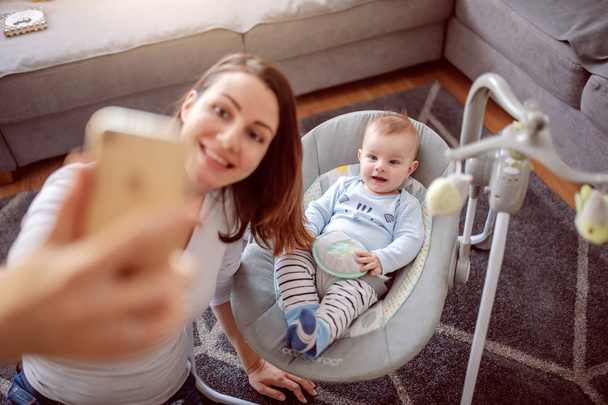 Vista superior de sonriente orgullosa madre caucásica tomando selfie con su adorable bebé. Bebé sentado en mecedora y sonriendo. Salón interior
. - Foto, Imagen