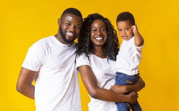 Jonge zwarte ouders met schattige zuigeling zoon over gele achtergrond - Foto, afbeelding
