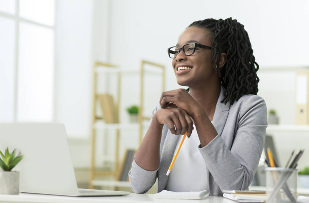 Χαμογελώντας μαύρη επιχειρηματίας κυρία κάθεται στο φορητό υπολογιστή στο χώρο εργασίας - Φωτογραφία, εικόνα