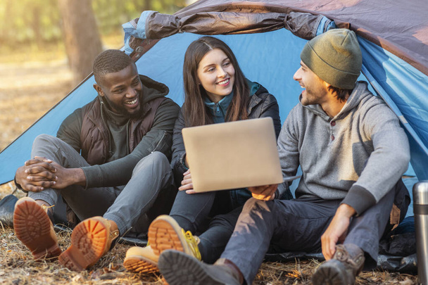 Trois amis heureux assis dans la tente de camping et en utilisant un ordinateur portable
 - Photo, image