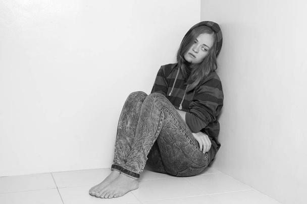 Νεαρή καυκάσια γυναίκα κάθεται στο πάτωμα, κλαίει, αναστατώνεται, κακοποιείται ή έχει κατάθλιψη. Μαύρο και άσπρο - Φωτογραφία, εικόνα