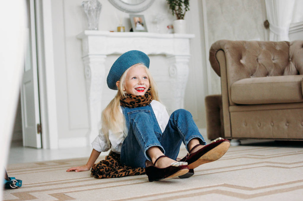 Cute fashion trendy fair hair little girl dressed in denim overalls, leopard scarf and blue beret with mom's accessories. Daughter imitates mom - Фото, зображення