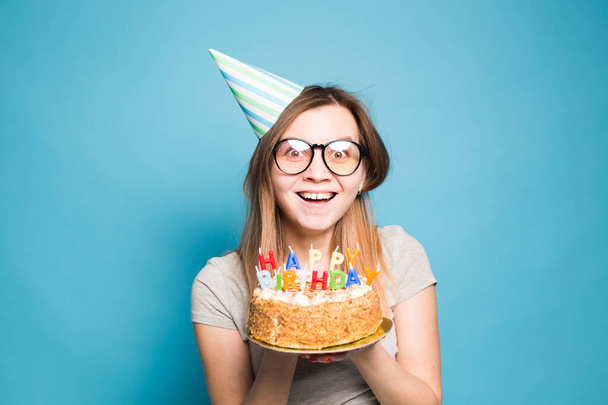 Nahaufnahme lustiges positives Mädchen mit Brille und grüßendem Papierhut, das auf blauem Hintergrund eine Geburtstagstorte in den Händen hält. - Foto, Bild