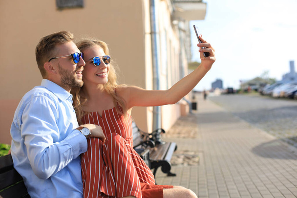 Romantic young couple in summer clothes smiling and taking selfie while sitting on bench in city street - Photo, image