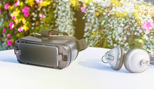 Equipo de auriculares de realidad virtual con auriculares inalámbricos de alta calidad para una experiencia virtual completa. Superficie de mesa blanca con flores de jardín en el fondo. Brillante día soleado
. - Foto, imagen