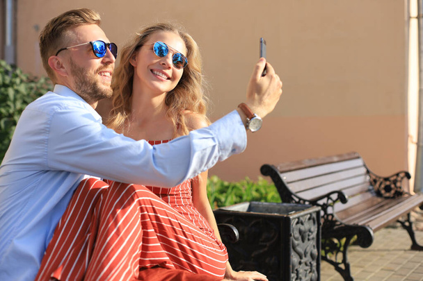 Romantic young couple in summer clothes smiling and taking selfie while sitting on bench in city street - Φωτογραφία, εικόνα