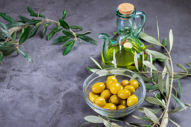 Aceitunas frescas en un recipiente de vidrio y una botella de aceite de oliva en el fondo oscuro
. - Foto, Imagen