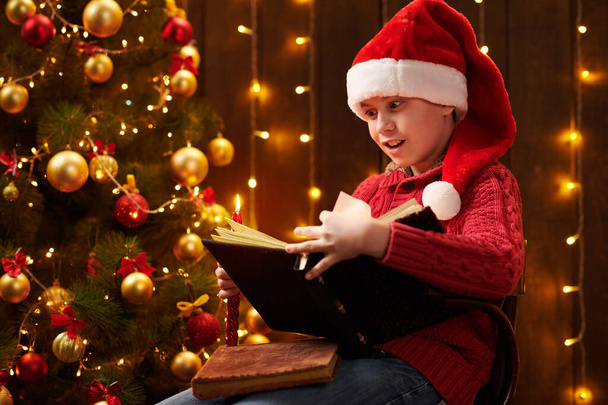 adolescent garçon lecture livre, assis à l'intérieur près décoré arbre de Noël avec des lumières, habillé comme Père Noël helper Joyeux Noël et Joyeuses Fêtes
! - Photo, image