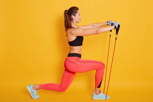 Caucasian woman coach in sporty short top and red gym leggings makes lunges by feet forward with fitness expander, attractive female athlete stretch legs with elastic expander in pilates studio. - Foto, afbeelding