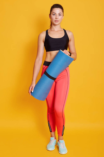 Toned young beautiful dak haired woman athlete wearing sportswear, standing and holding blue fitness mat, looking directly at camera, fit female after yoga session or training. Sport concept. - Fotó, kép