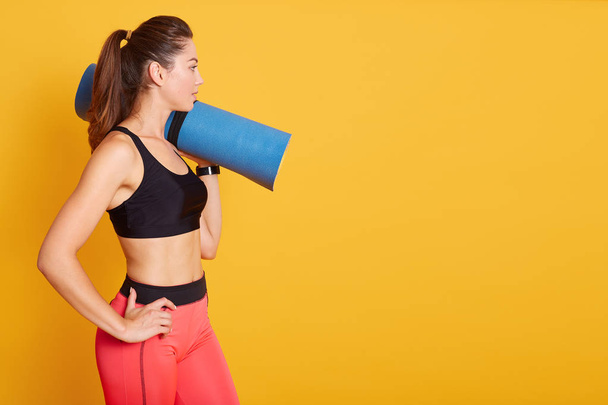 Side wiev of young beautiful Caucasian woman standing and holding blue yoga mat over shoulder, being ready for exercising in gym, wearing stylish sportwear. Copy space for advertisment. Sport concept. - Valokuva, kuva