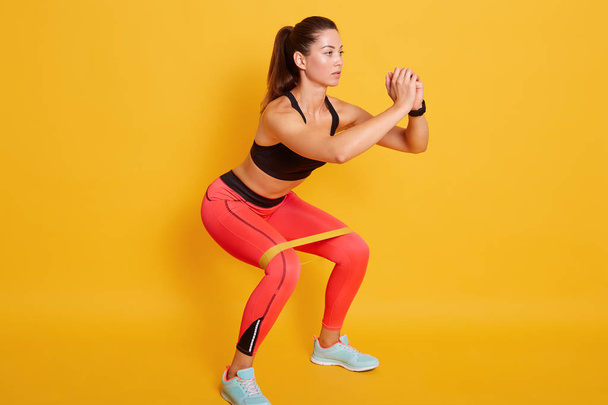 Studio shot of sporty woman squatting, doing sit ups with resistance band. Photo of Caucasian woman in fashionable sportswear isolated over yellow background. Strength and motivation concept. - Photo, Image