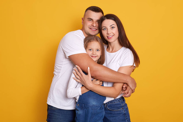 Horizontal shot of beautiful young family hugging, looking directly at camera and smiling while standing against yellow studio wall, people dress casually, expressing happyness. Relationship concept - Photo, Image