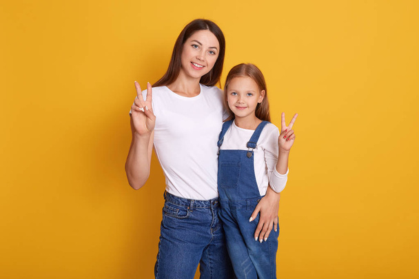 Close up portrait of happy woman showing peace sign with her charming daughter, woman wearing white t shirt and jeans, kid dresses ovealls, expresses happyness, isolated over yellow studio background. - Photo, Image