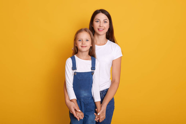 Horizontal shot of mother with daughter, being happy spending together, female dresses white t shirt and jeans, cute little girl wearing overalls, family look directly at camera. People concept. - Foto, Imagen