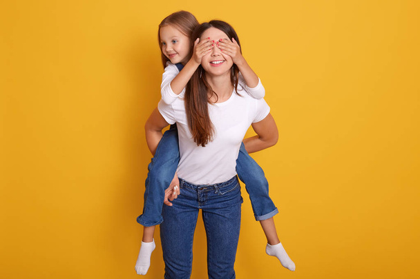 Indooor shot of happy mother piggybacking daughter while cute kid closing her eyes, posing isolated over yellow studio background, happy woman having fun with charming girl at home. Family concept. - Fotó, kép