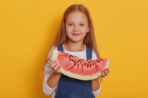 Indoor shot of little charming girl with portion of sweet watermelon in her hands, charming blonde modelposing isolated over yellow studio background, child wearing white shirt and overalls. - Foto, imagen