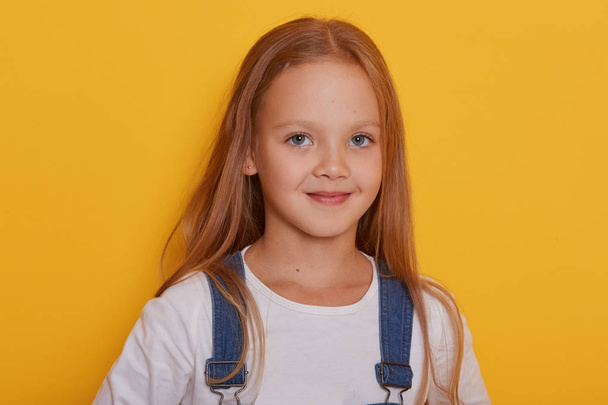 Indoor shot of beautiful little girl with long blonde hair posing isolated over yellow background, looking at camera, charming kid wearing casual outfit, being glad to be photographed in studio. - Foto, immagini