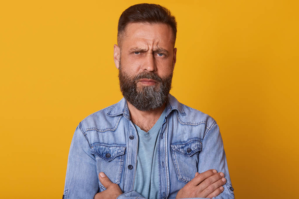 Close up portrait of fashionable middle aged man with beard and trendy haircut wearing denim jacket, posing isolated against yellow studio wall background with serious or angry facial expression. - Valokuva, kuva