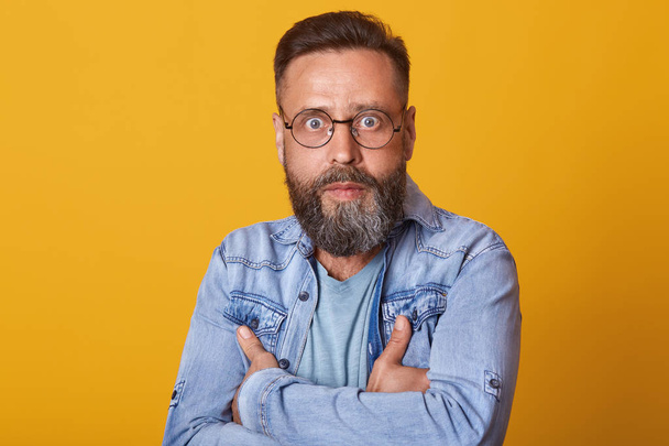 Portrait of shocked middle aged man in full disbelief. Hipster male with beard wears denim jacket posing isolated over yelow studio background. Astonished face expression, human emotion, body language - Fotoğraf, Görsel