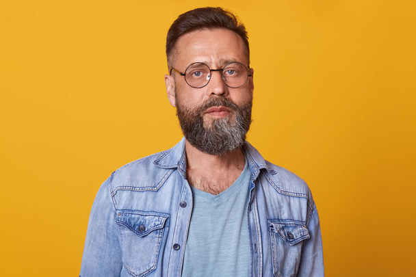 Photo of bearded man with folded hands, standing and looking directly at camera with concentrated facial expression, being in good mood, dressed stylish denim jacket and gray t shirt. People concept. - Photo, image